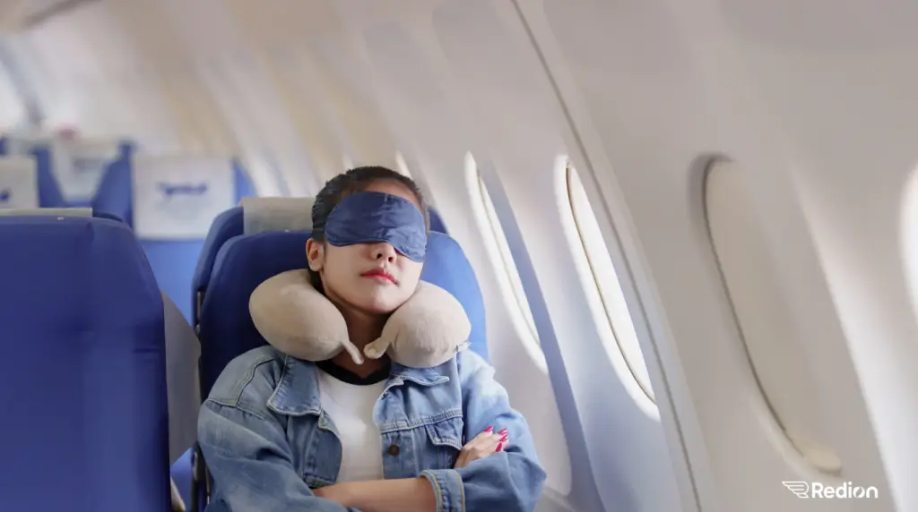 jeune fille avec un masque sur les yeux et qui se repose dans un avion, assise à côté d'un hublot.