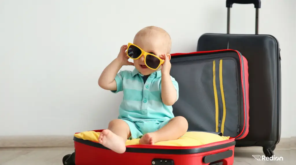 bébé dans une valise ouverte et qui joue avec de grosses lunettes de soleil jaunes.